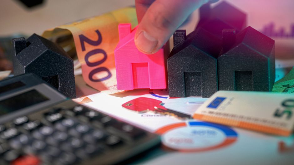 A close-up view of a businessman's hand holding a small pink house-shaped model made of plastic, positioned among three larger black house-shaped models with textured surfaces that resemble speckled s